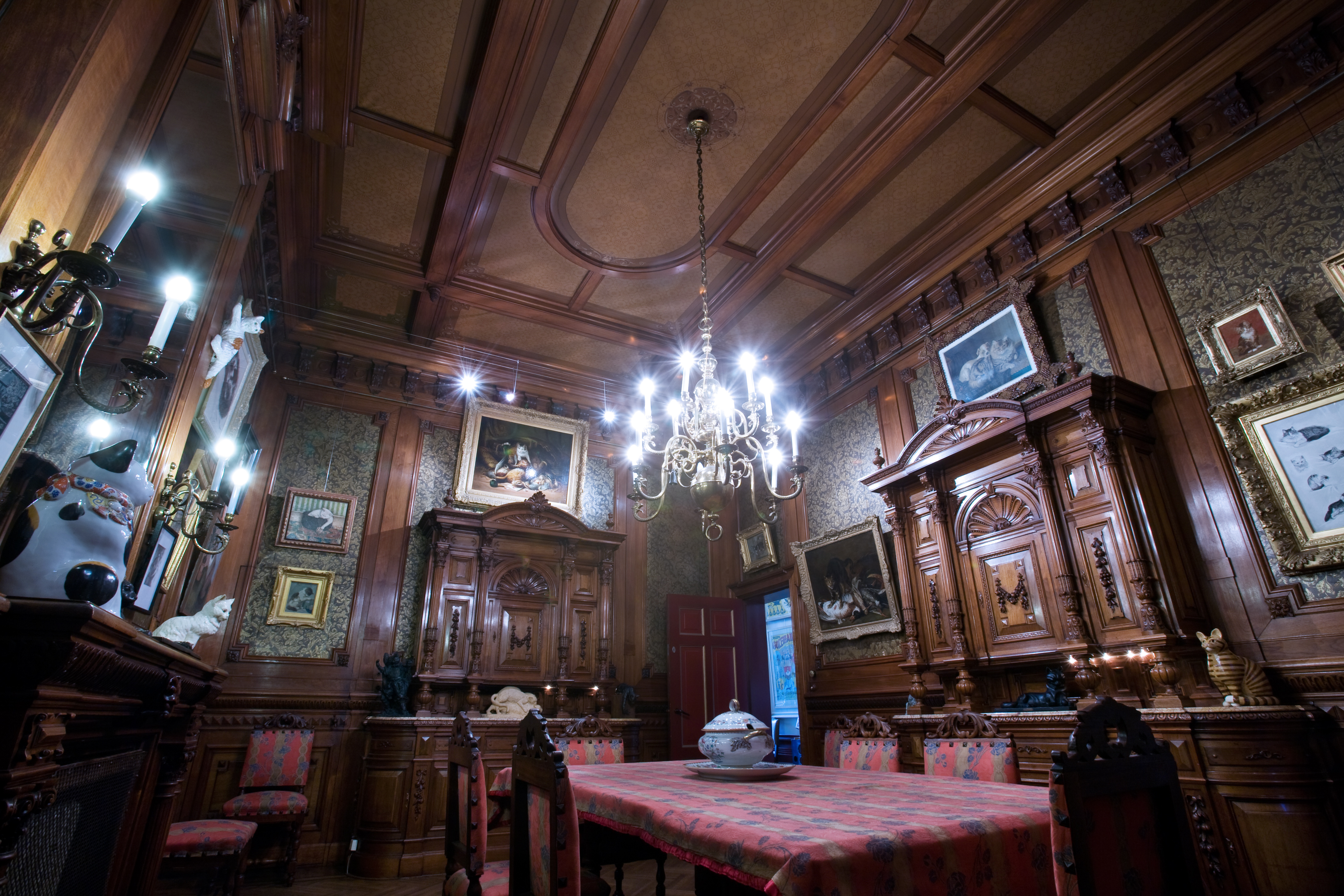 Sierlijke museumzaal met donkerhouten lambrisering, kroonluchter, ingelijste schilderijen en een kattenbeeldje op een tafel met rood patroon. 
