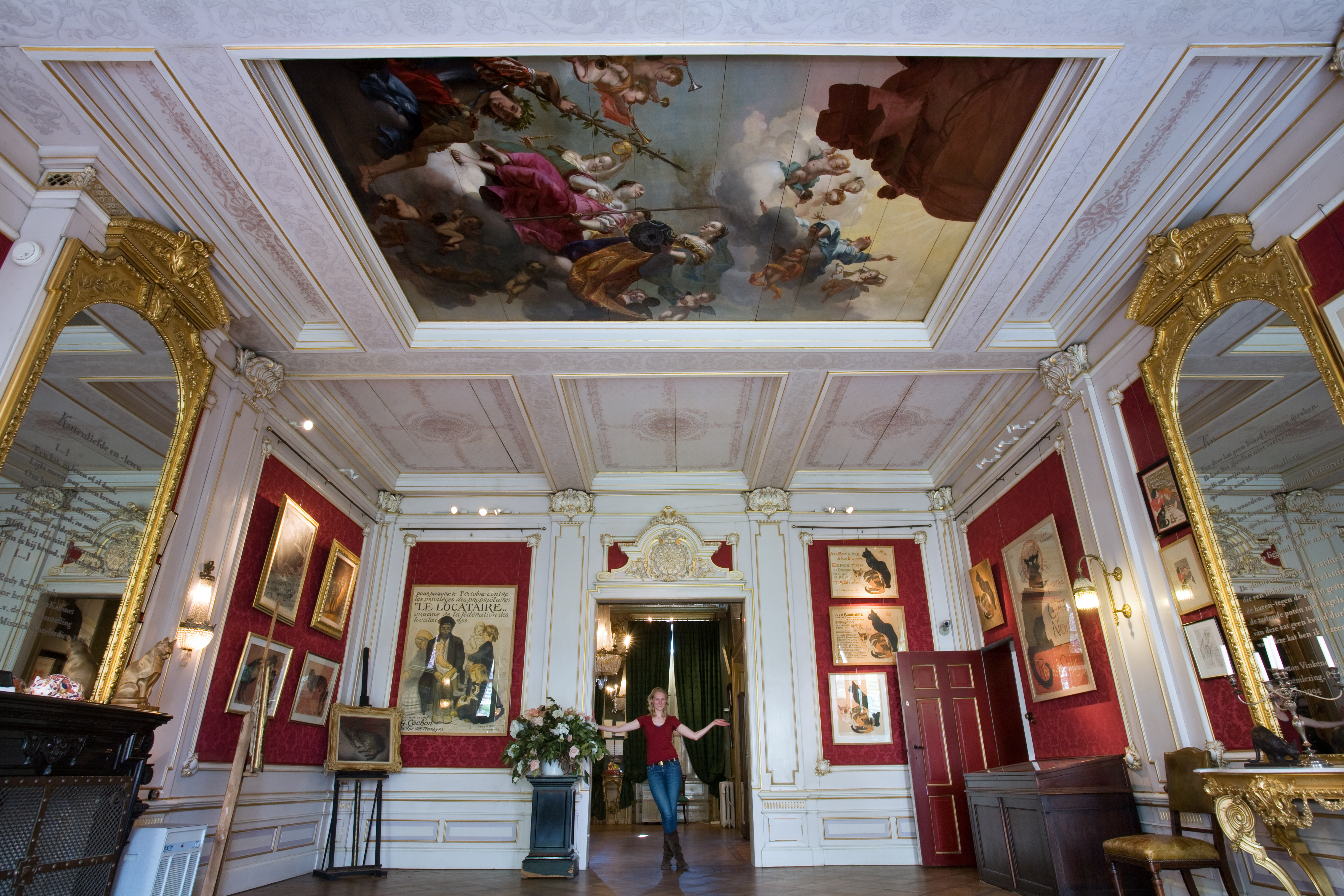 Sierlijk interieur van KattenKabinet met een groot plafondfresco, rode muren, gouden spiegels en een persoon die in het midden staat