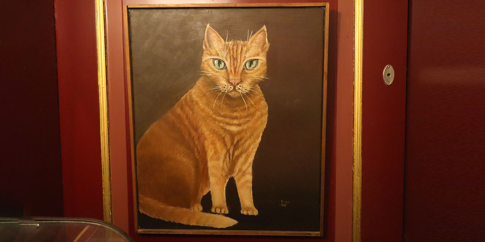 Framed oil painting of an orange tabby cat with green eyes displayed on a red wall in a museum setting