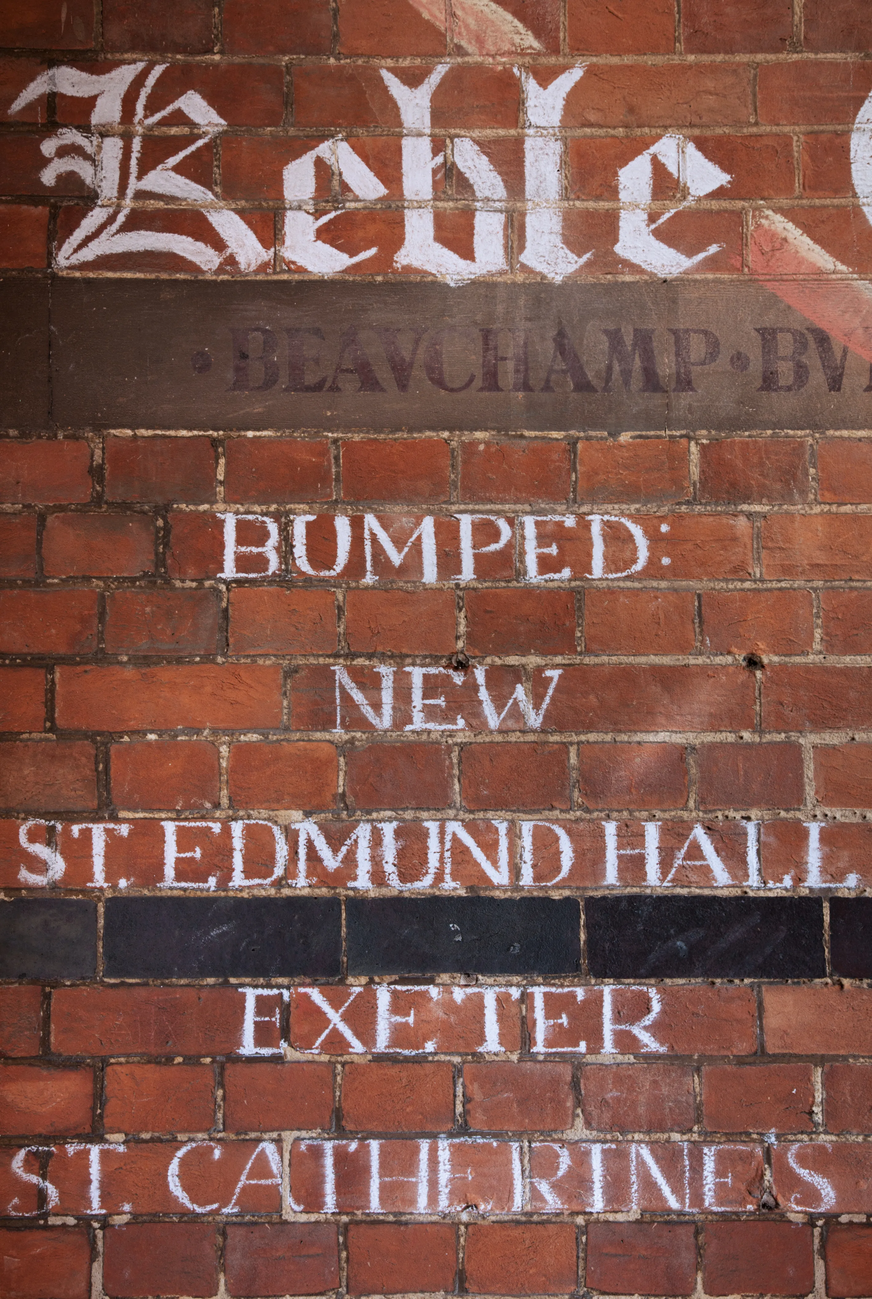 Zbliżenie na ścianę z czerwonej cegły w Keble College z białym napisem gotycką czcionką: 'Keble', 'BUMPED:', 'NEW', 'ST. EDMUND HALL', 'EXETER' oraz 'ST. CATHERINE'S'