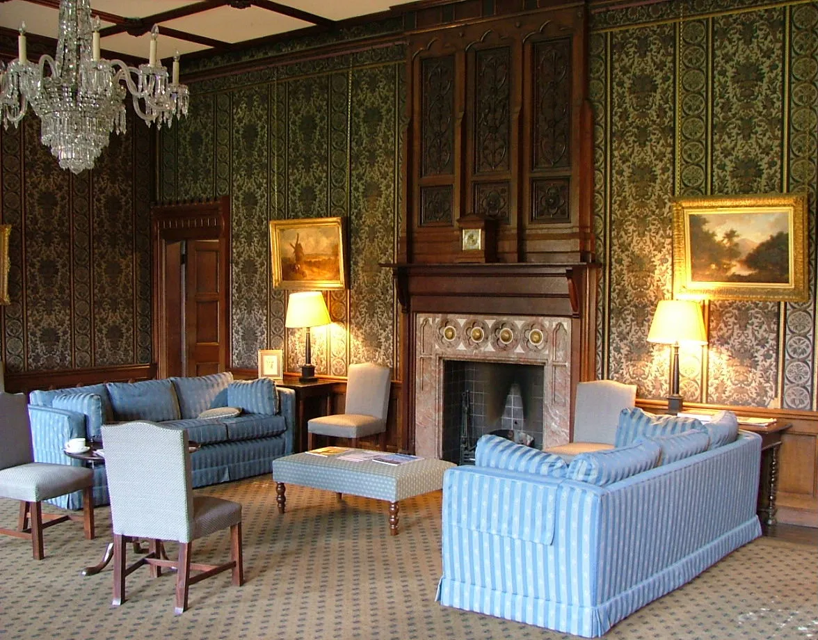 Formele zitkamer met blauwgestreepte banken en fauteuils opgesteld rondom een grote houten schouw, met groen behang met patronen, een kristallen kroonluchter en ingelijste schilderijen aan de muren.