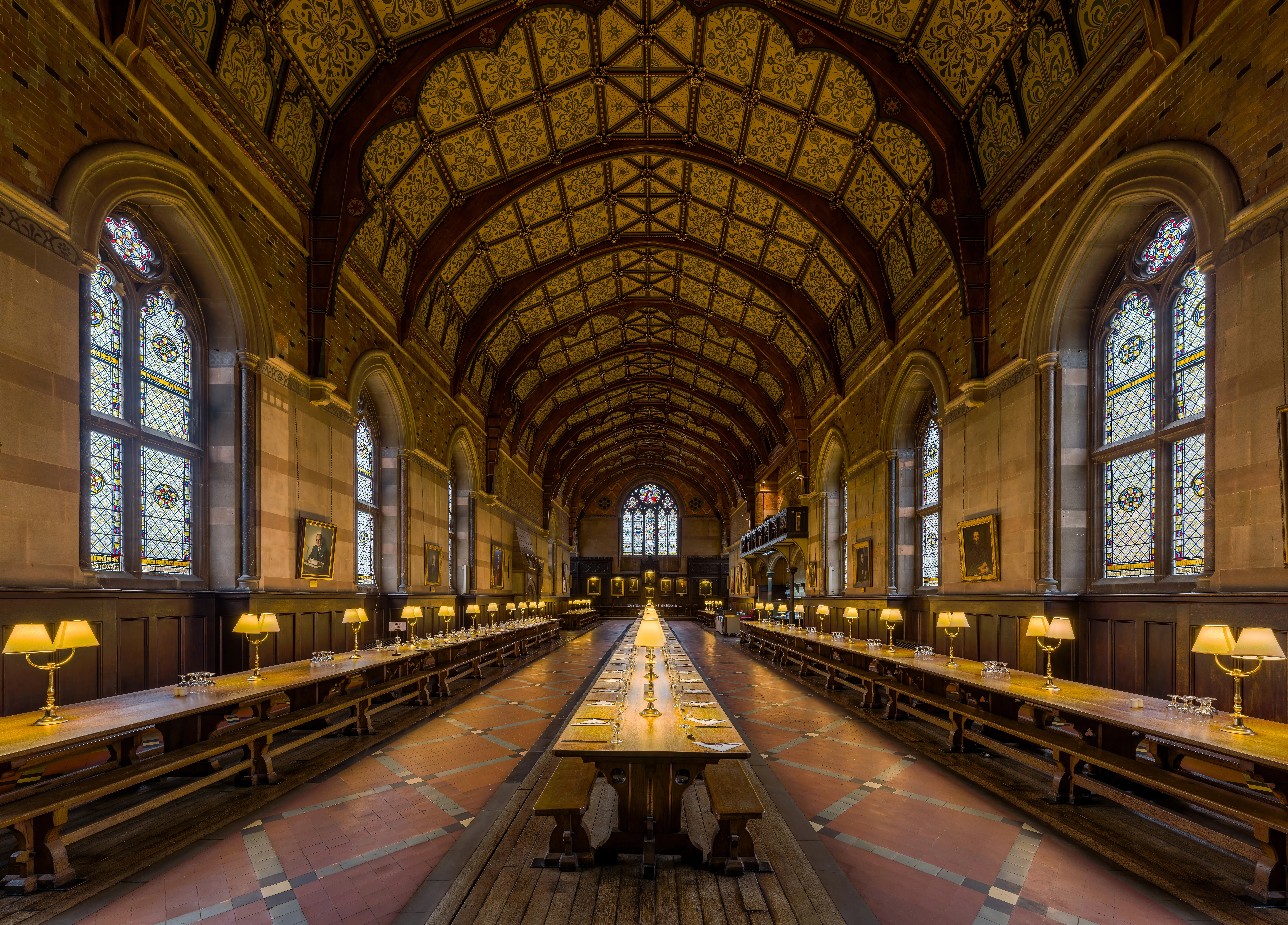 Lange, rijkelijk versierde eetzaal met een uitbundig gedecoreerd gewelfd plafond, lange houten tafels en banken, glas-in-loodramen en warme tafellampen in Keble College, Oxford
