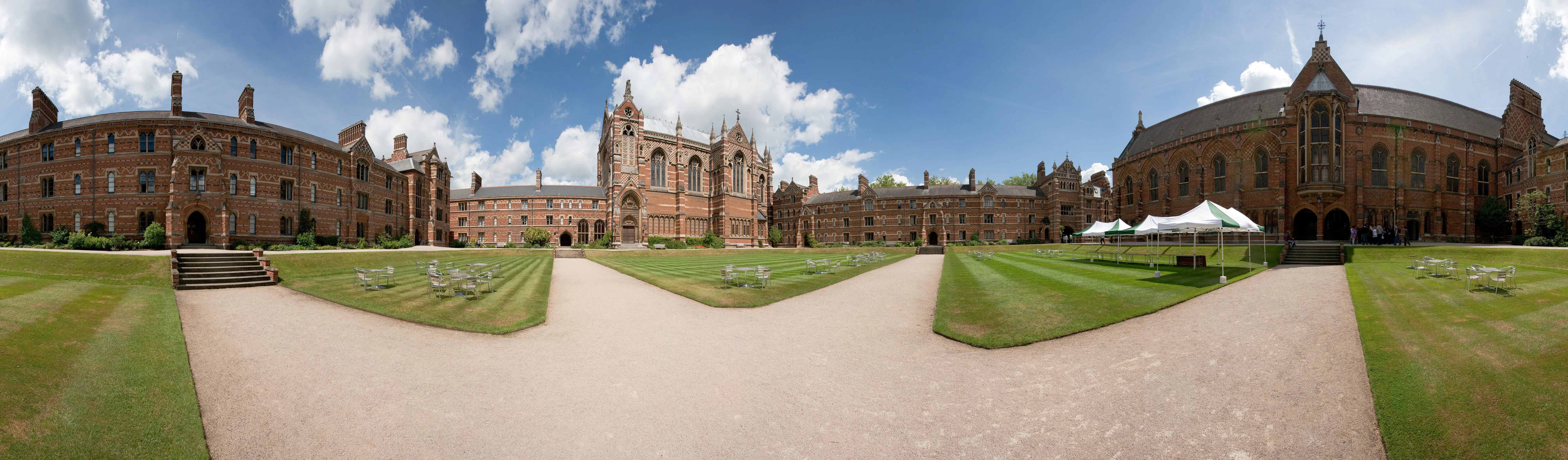 Panoramisch uitzicht op de binnenplaats van Keble College met rood- en geelgekleurde bakstenen gebouwen, een centrale kapel en groene gazons onder een blauwe lucht met wolken.