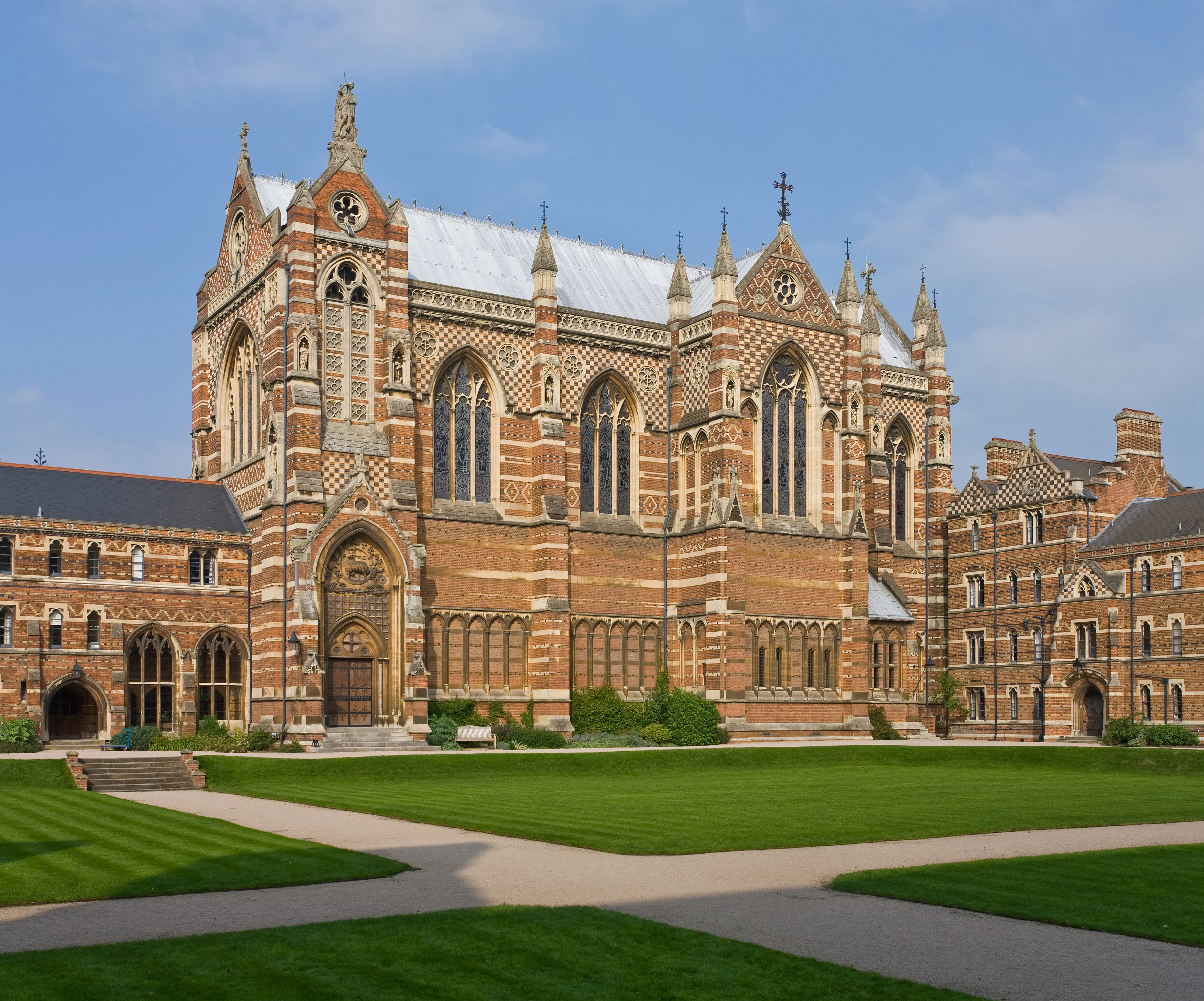 De polychrome rood- en geelbakstenen gevel van de Keble College Chapel met gotische boogramen, stenen pinakels en een verzorgd groen gazon op de voorgrond onder een blauwe lucht.