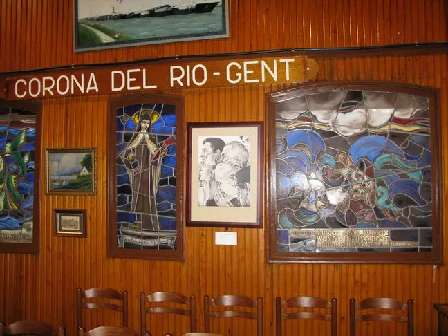 Pared interior revestida de madera con paneles de vidrieras, obras de arte enmarcadas y un letrero que dice CORONA DEL RIO - GENT sobre sillas de madera