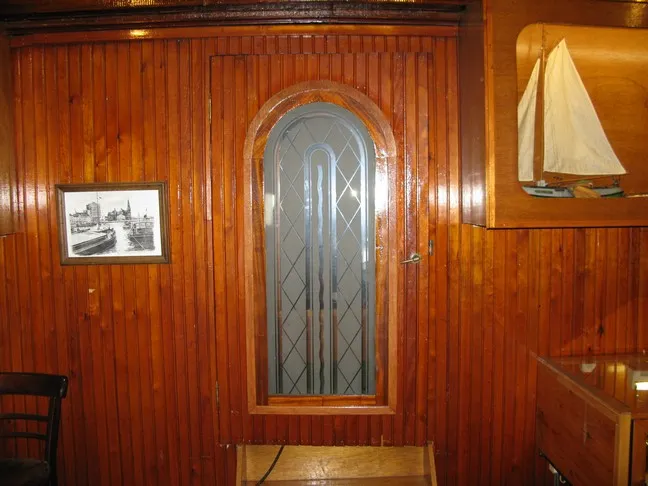 Habitación interior con paneles de madera que cuenta con una ventana arqueada de vidrio esmerilado, una fotografía en blanco y negro enmarcada de un barco en la pared y un modelo de velero expuesto en un estante de madera.
