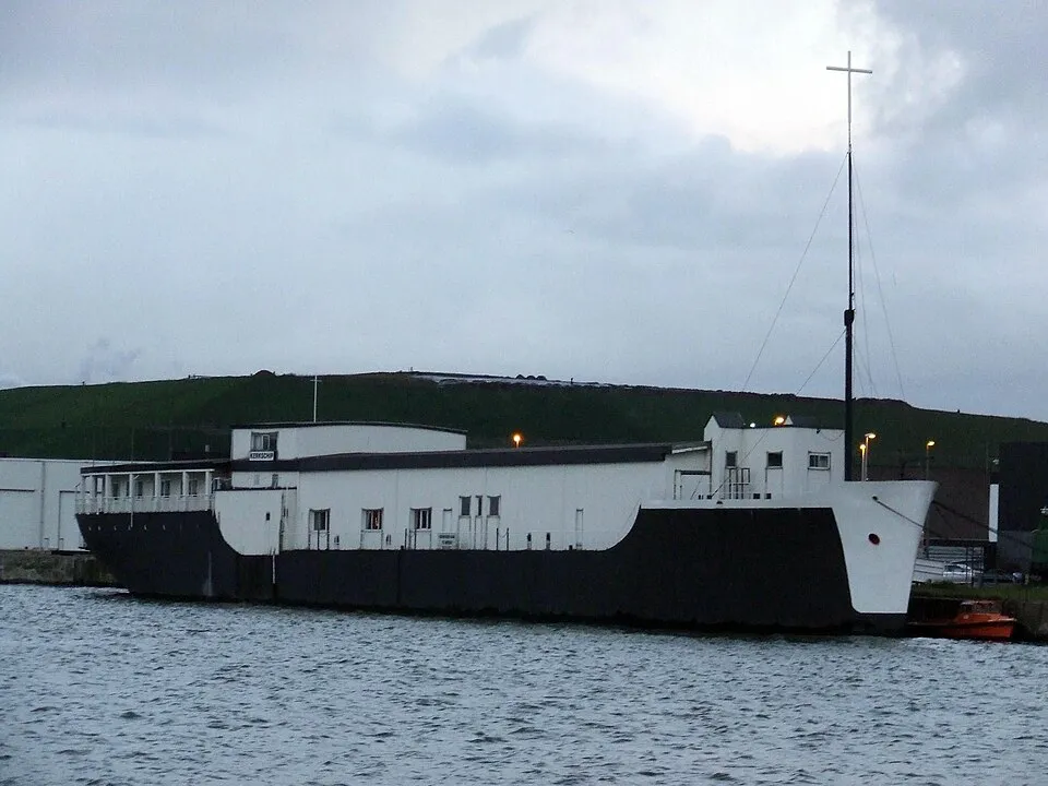 Un gran barco-iglesia en blanco y negro con una cruz alta en su mástil flotando en el agua, con una colina verde de fondo bajo un cielo nublado