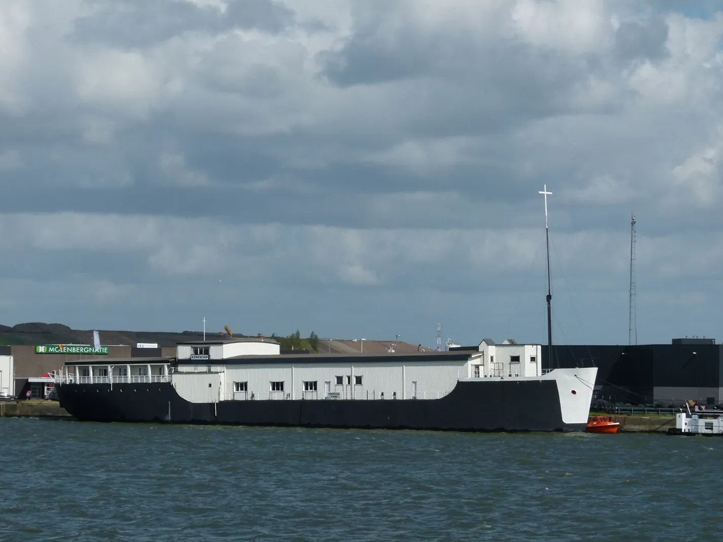 Iglesia barco de hormigón Sint-Jozef atracada en un canal, que presenta una superestructura blanca, casco negro y un alto mástil con cruz sobre un cielo nublado