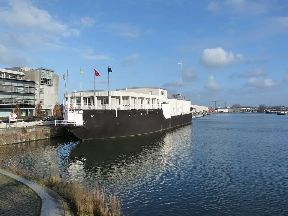 Iglesia barco de hormigón en blanco y negro atracada en un canal con banderas ondeando en la parte superior, junto a un edificio moderno bajo un cielo azul