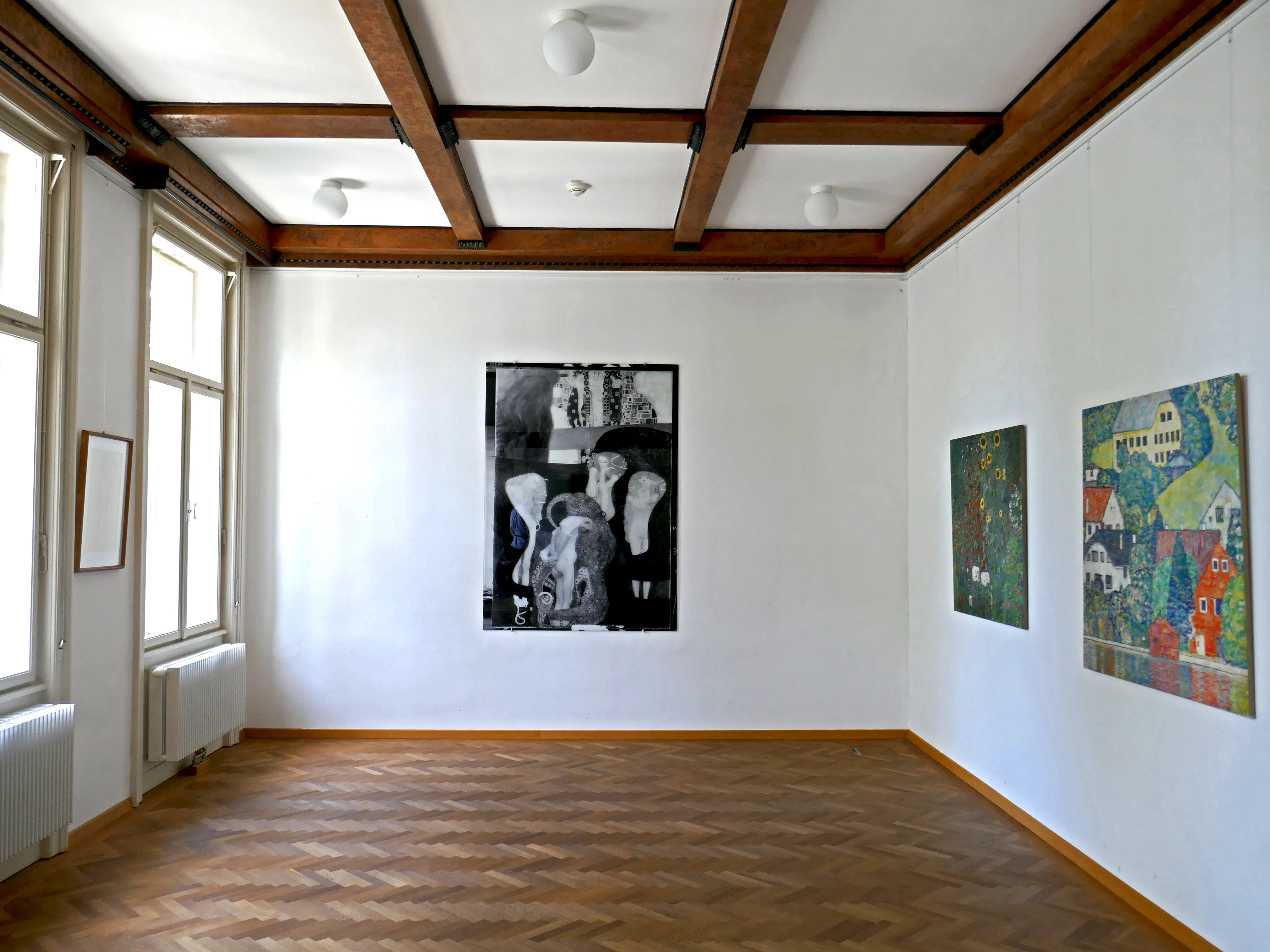 Interior of a gallery room with wooden beams, parquet flooring, and three artworks on white walls