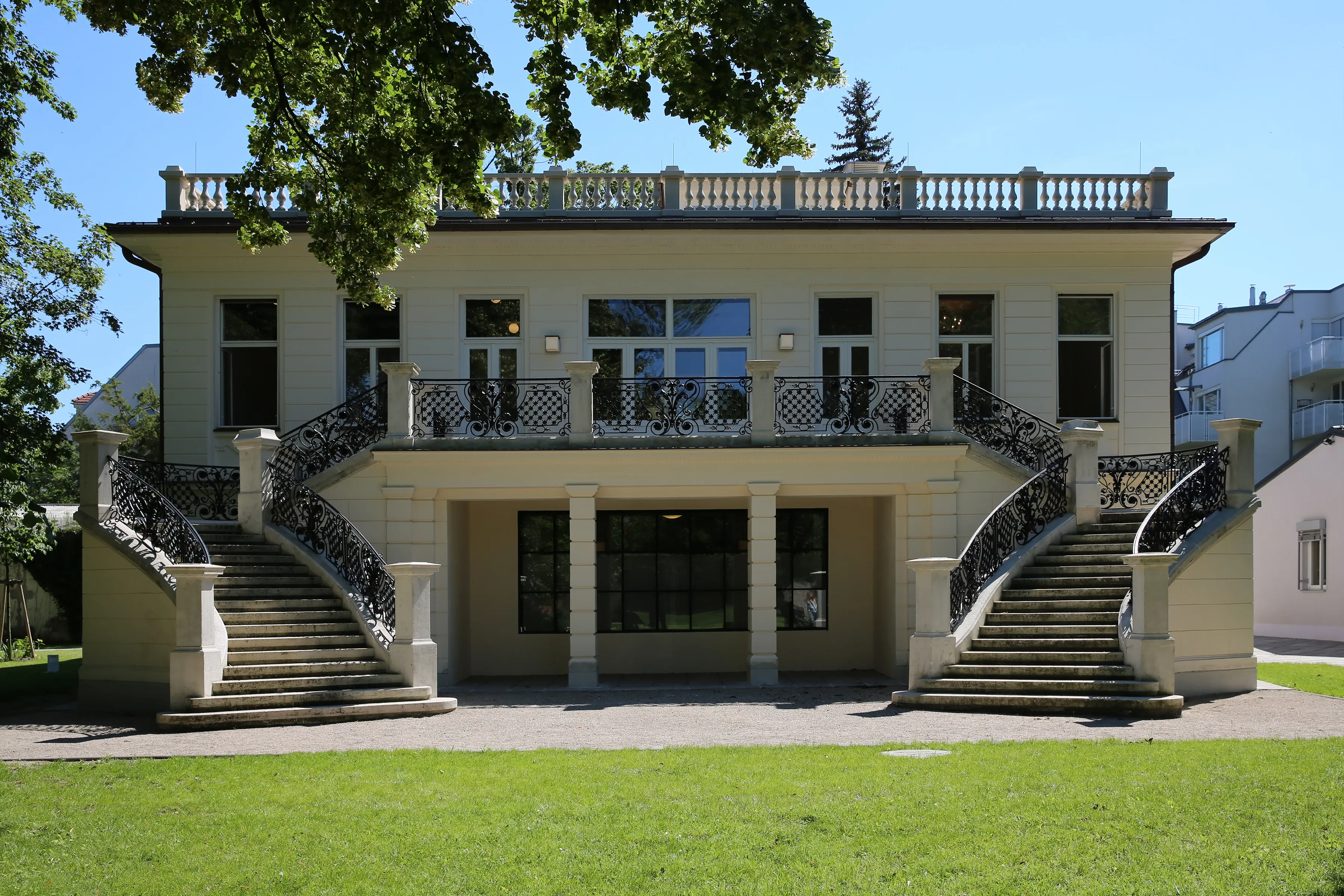 A two-story building with a cream facade and double staircases leading to a central balcony, surrounded by green grass and trees under a clear blue sky