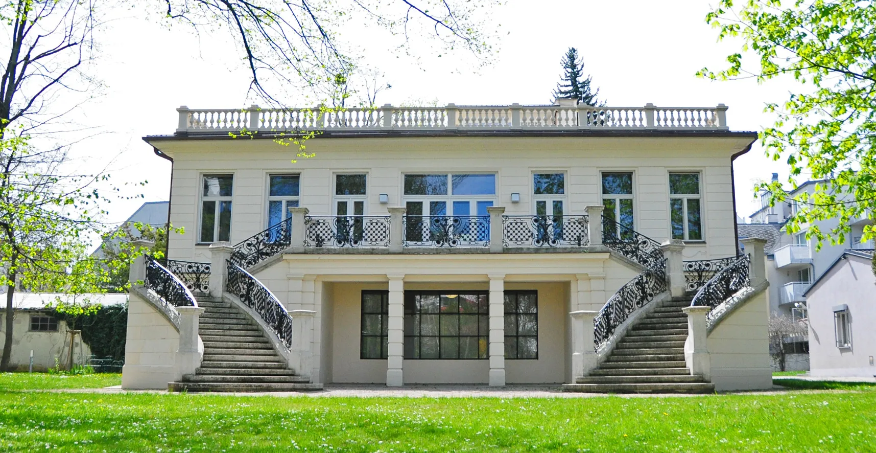 White two-story building with a central balcony and dual staircases surrounded by green grass and trees