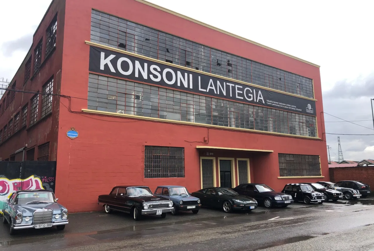 Edificio industrial de ladrillo rojo con grandes letras KONSONI LANTEGIA en la fachada y coches antiguos aparcados delante bajo un cielo nublado