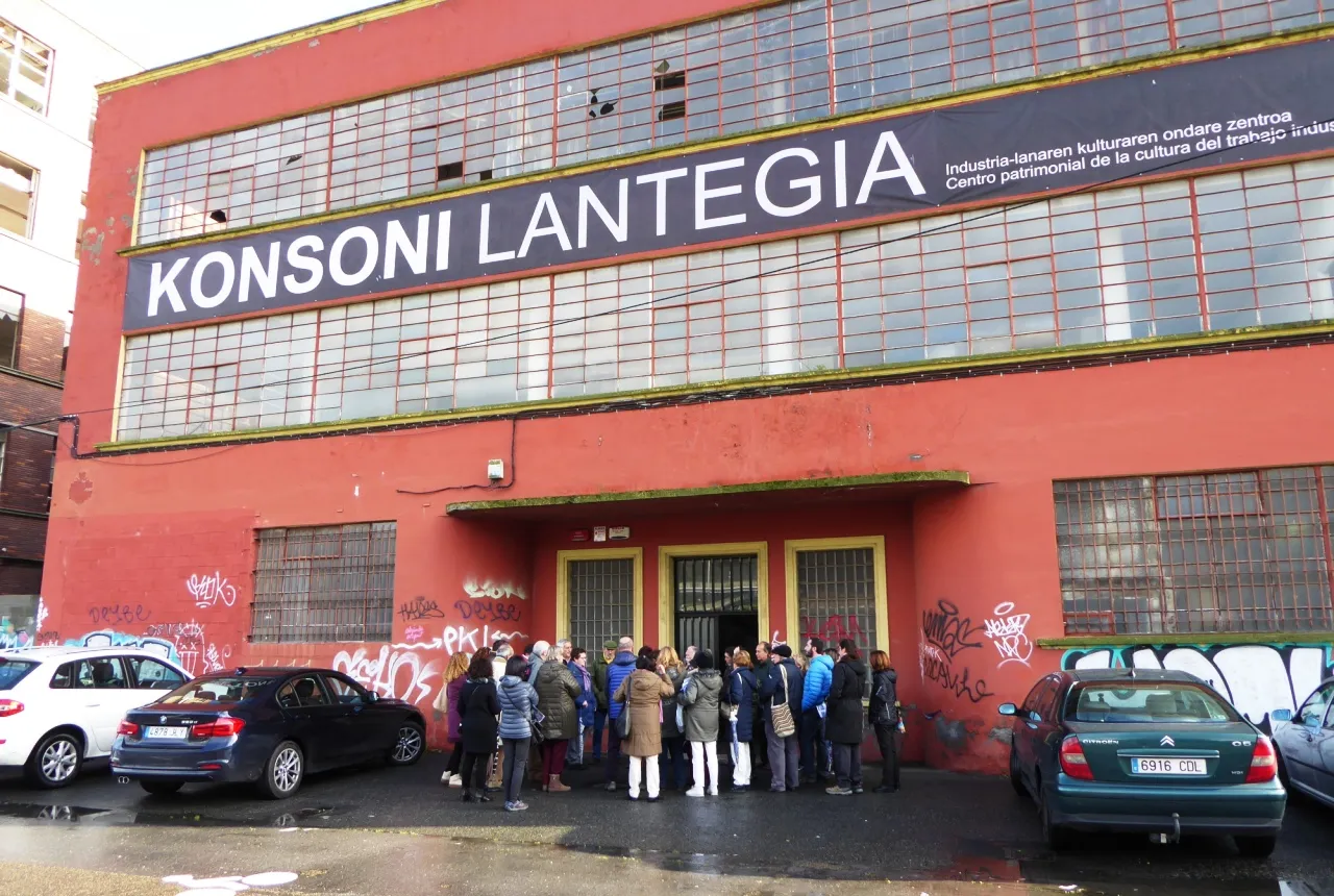 Fachada de un edificio industrial rojo con un gran cartel "KONSONI LANTEGIA" sobre la entrada, personas reunidas afuera y coches aparcados delante