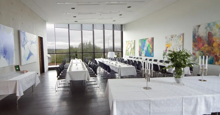 Bright interior hall with long tables covered in white tablecloths, rows of chairs, large floor-to-ceiling windows, and colorful abstract paintings on the walls.