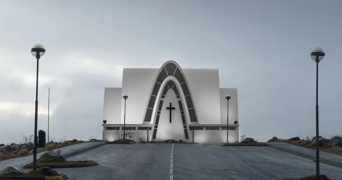 White modernist church building with large intersecting arches and a central cross, viewed from a paved road with tall lamp posts on either side under an overcast sky