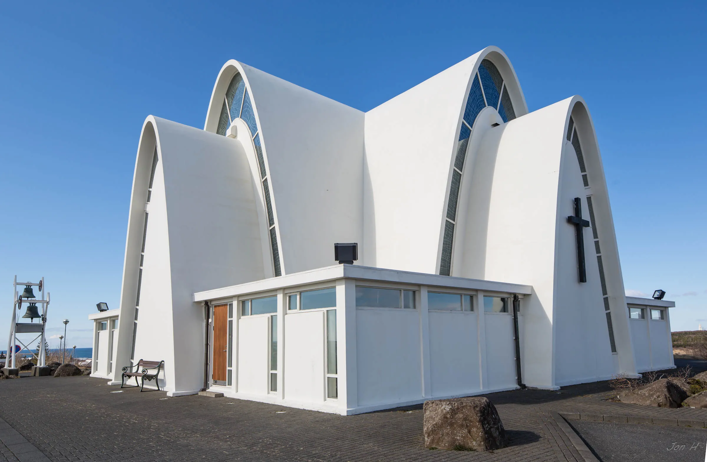 White modernist church with distinctive intersecting arched rooflines and a black cross on the facade under a clear blue sky
