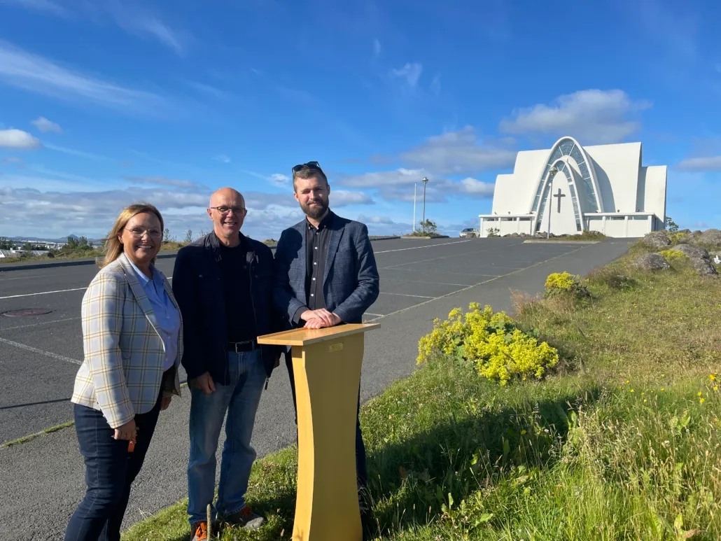 Three people standing at a wooden podium outdoors with the white modernist Kópavogskirkja church visible in the background under a blue sky