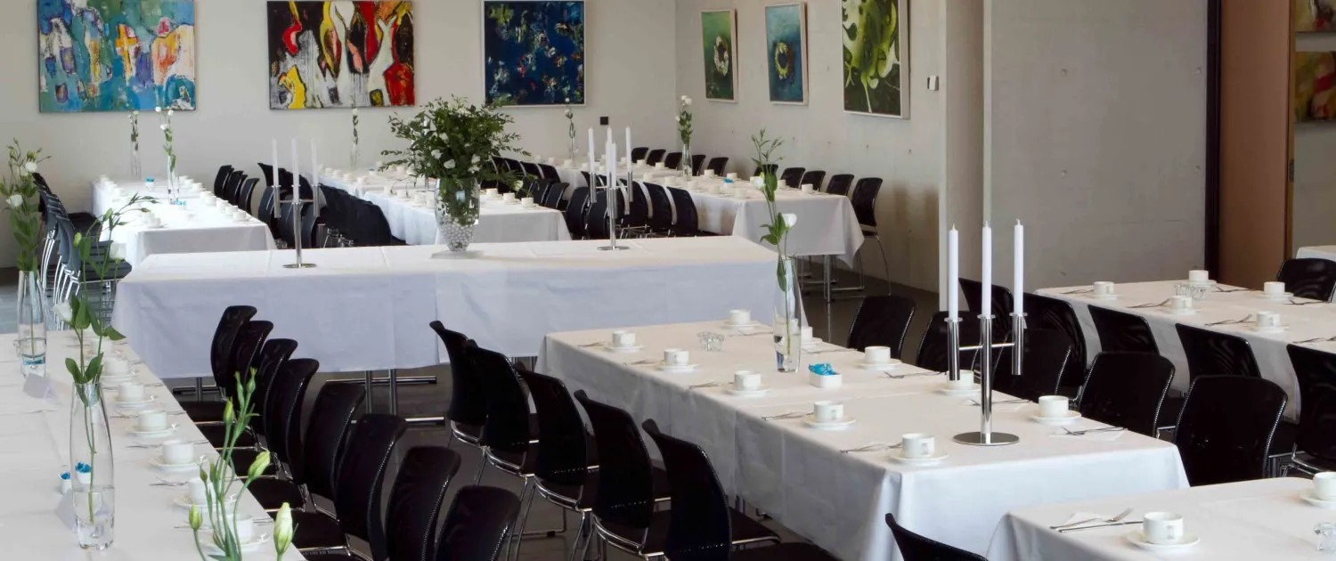 A long hall set for a banquet with white-clothed tables, black chairs, tall candle holders, and colorful paintings on the walls.
