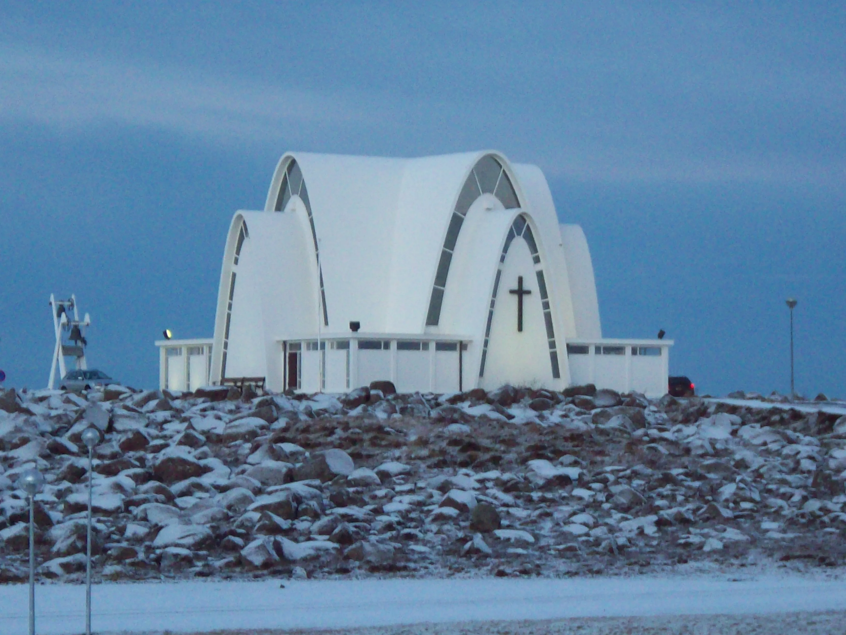 White modernist church with intersecting arched rooflines and a cross on the facade, sitting atop a rocky snow-covered hill under an overcast sky