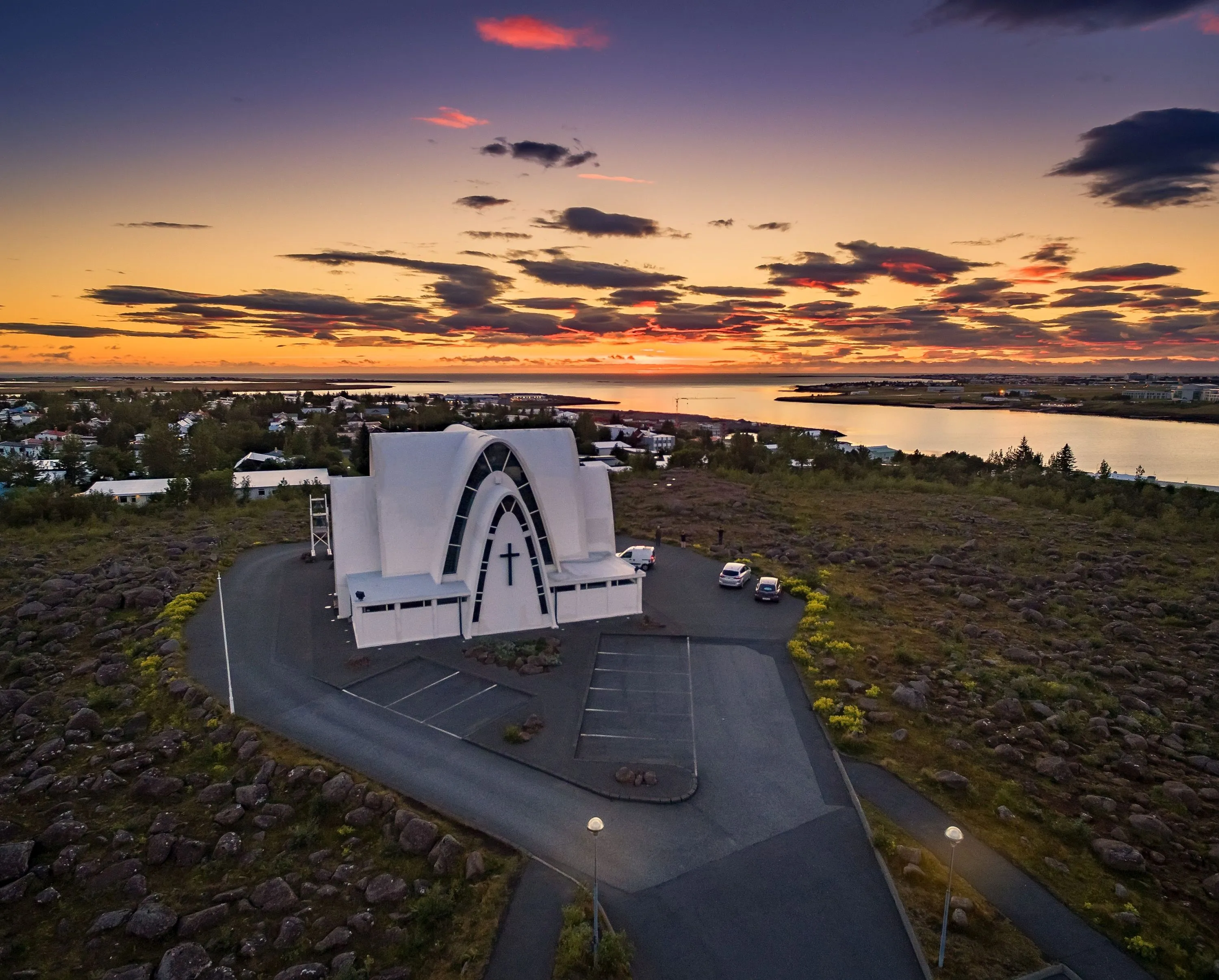 Aerial view of Kópavogskirkja, a white modernist church with intersecting arches, on a lava field hilltop at sunset with colorful orange and purple sky and water in the background