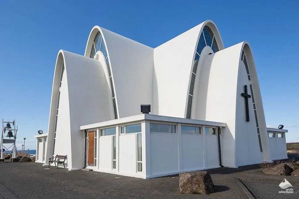 White modernist church with tall intersecting arched forms, large stained glass windows, and a black cross on the facade under a clear blue sky
