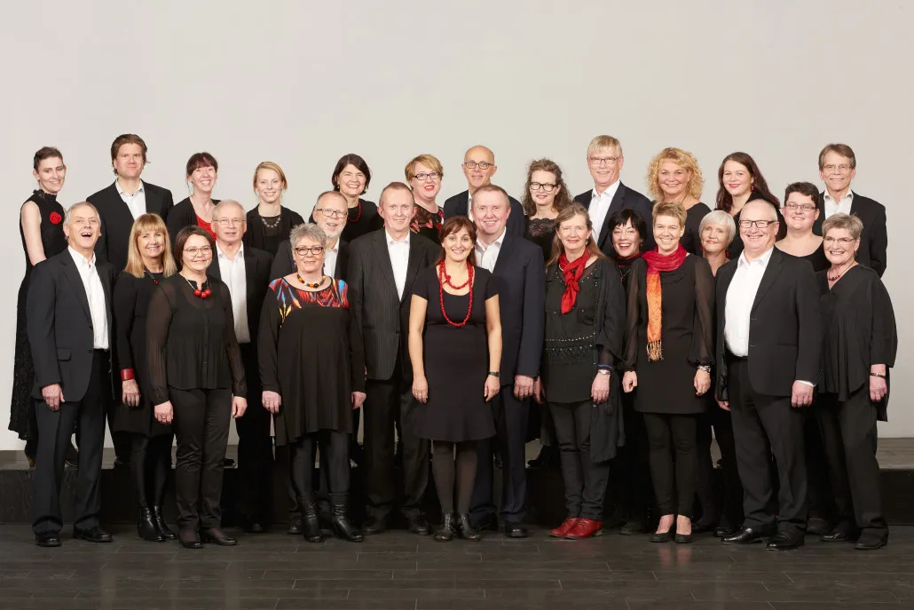 Duża grupa chóralna składająca się z około 25 osób w formalnych czarnych strojach z czerwonymi szalami i naszyjnikami, stojąca w rzędach na scenie na tle jednolitego jasnego tła