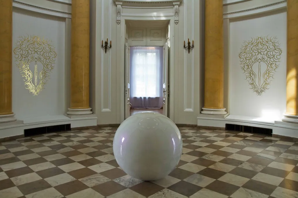 Een witte sferische sculptuur in een statige hal met een dambordvloer en sierlijke wanddecoraties