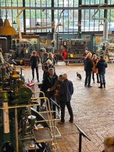 Interior de museo industrial con suelo de ladrillo y visitantes examinando maquinaria pesada bajo ventanas altas