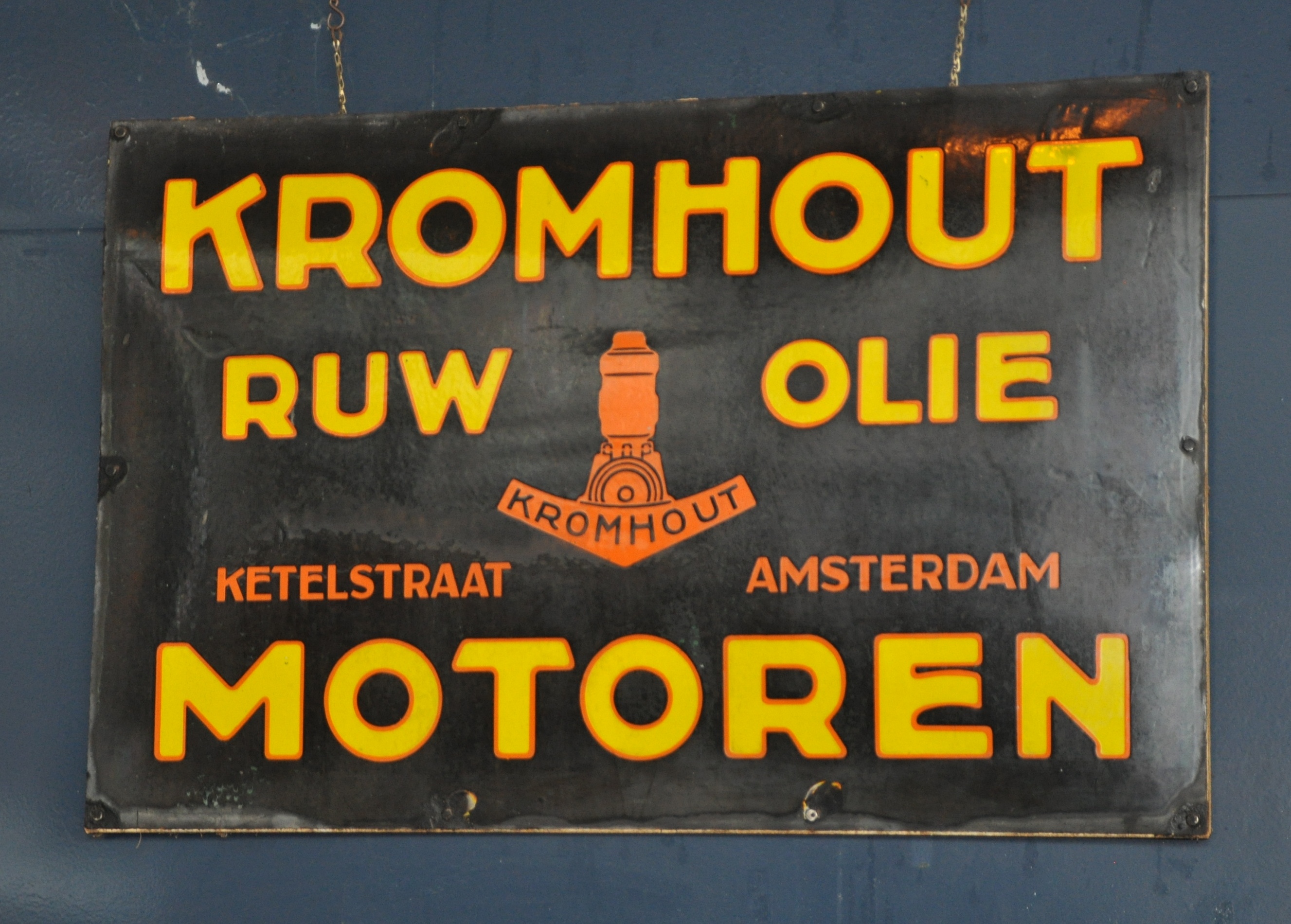 Letrero negro vintage con texto amarillo 'KROMHOUT RUW OLIE KETELSTRAAT AMSTERDAM MOTOREN' y un emblema naranja de motor de barco.