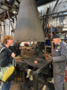 Dos personas conversan cerca de un gran horno industrial entre herramientas y maquinaria en un taller.