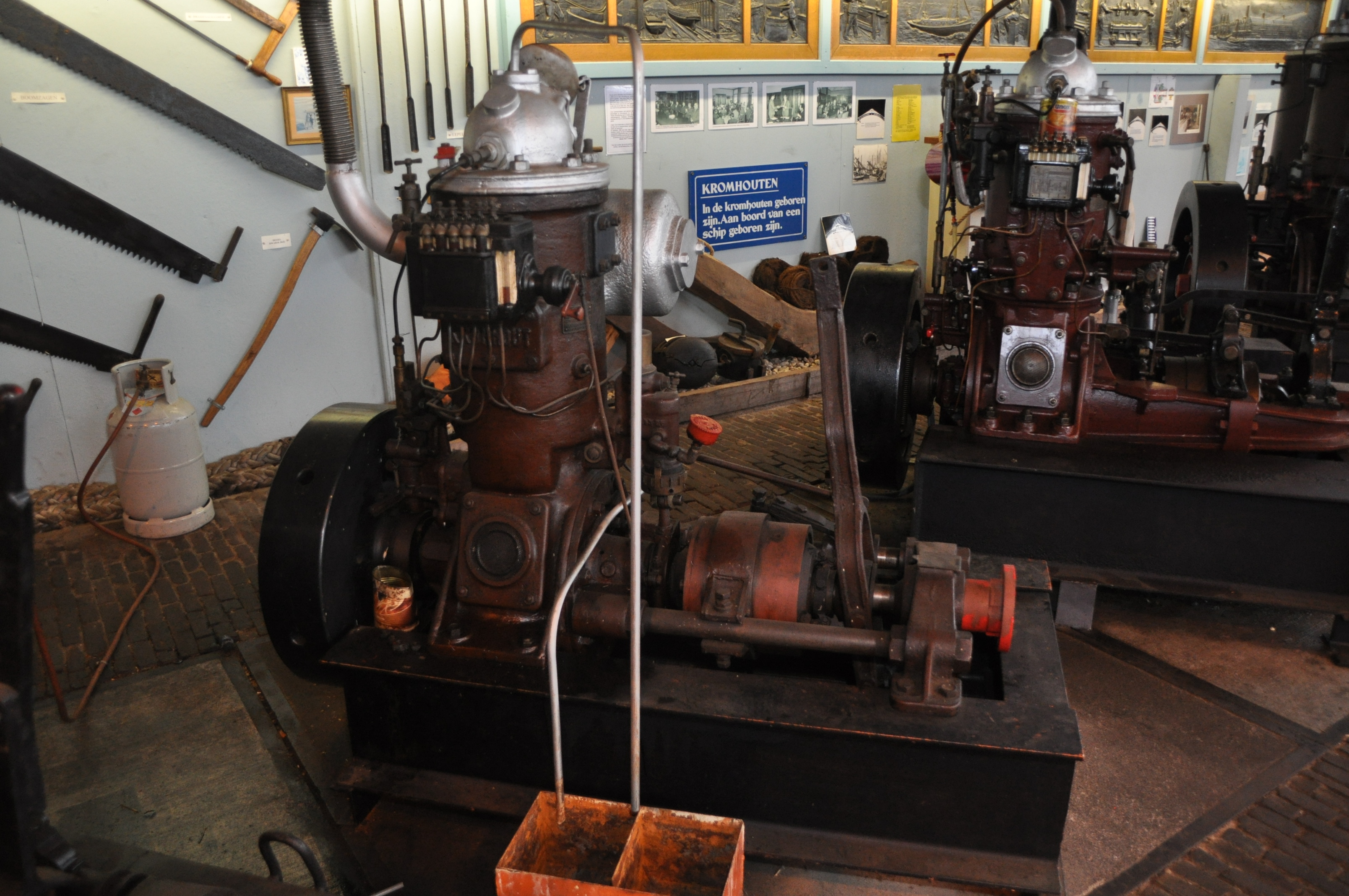 Large vintage ship engines on display in a museum workshop with concrete flooring and an informational sign on the wall.