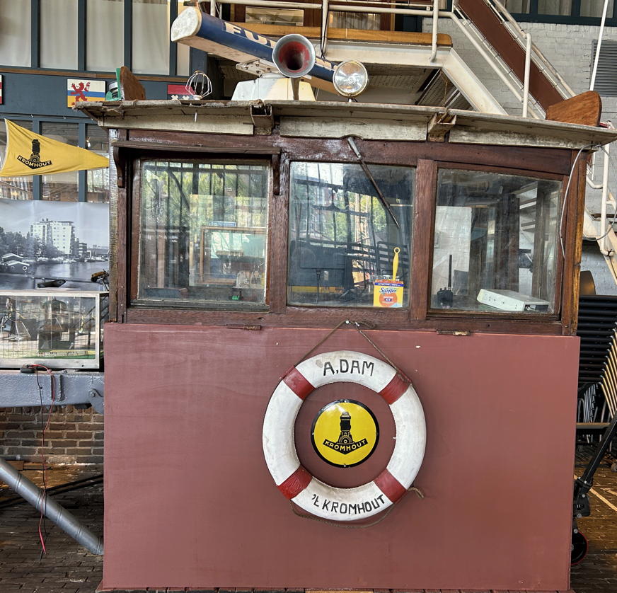 Timonera de barco de época con una] [boya salvavidas roja y blanca etiquetada 'A.DAM KROMHOUT', expuesta entre exhibiciones industriales en un entorno de museo