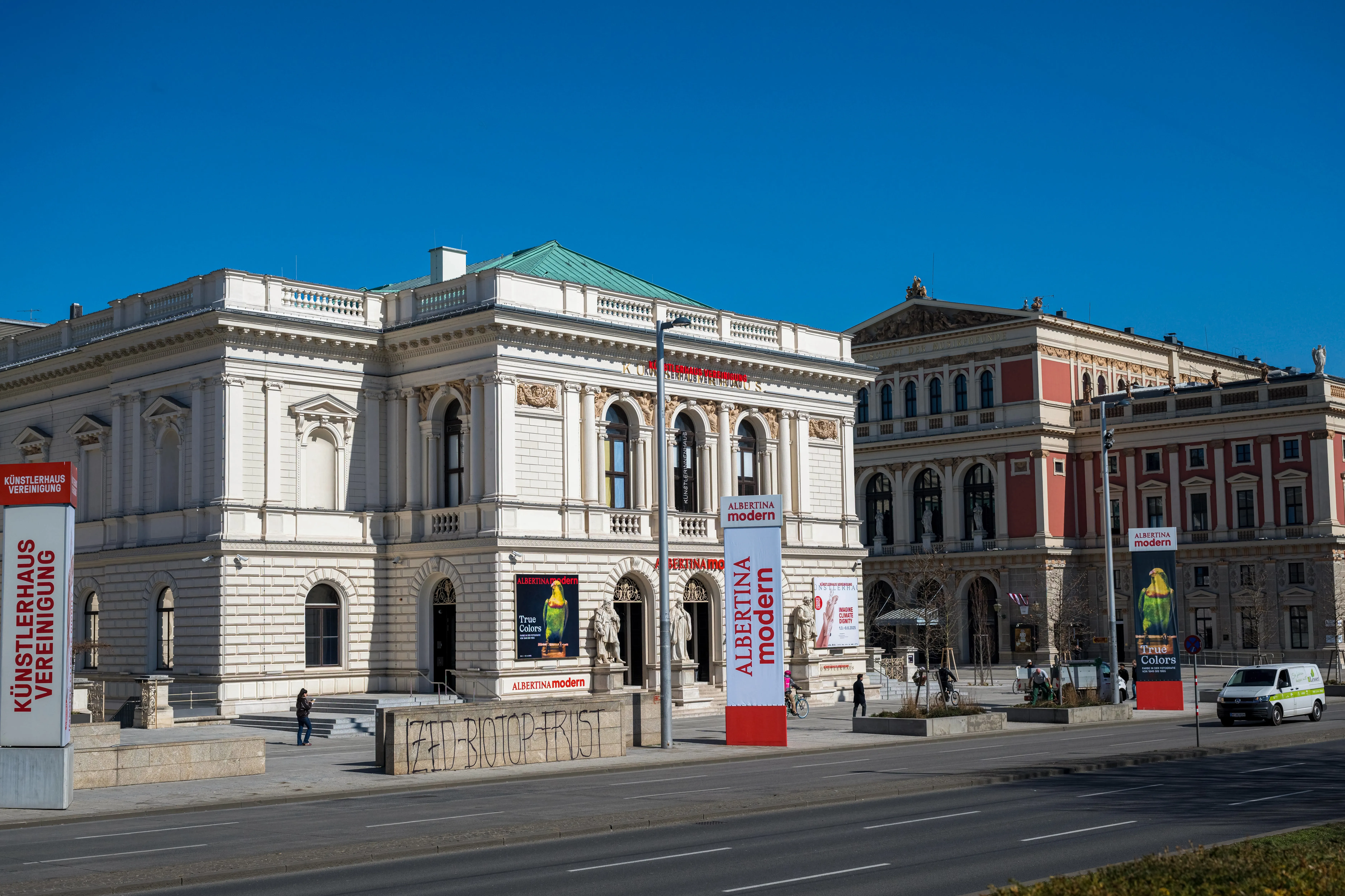 Klassiek wit gebouw met boogramen en een groen dak, met rode spandoeken en bewegwijzering voor Künstlerhaus Wien op een zonnige dag.