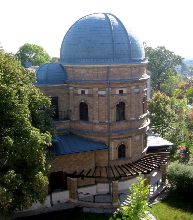 Un edificio observatorio histórico con una cúpula azul y fachada de ladrillo, rodeado de árboles y vegetación.