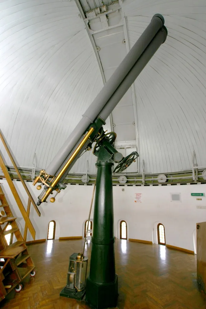 Telescopio grande dentro de una sala de observatorio abovedada con pisos de madera y ventanas arqueadas