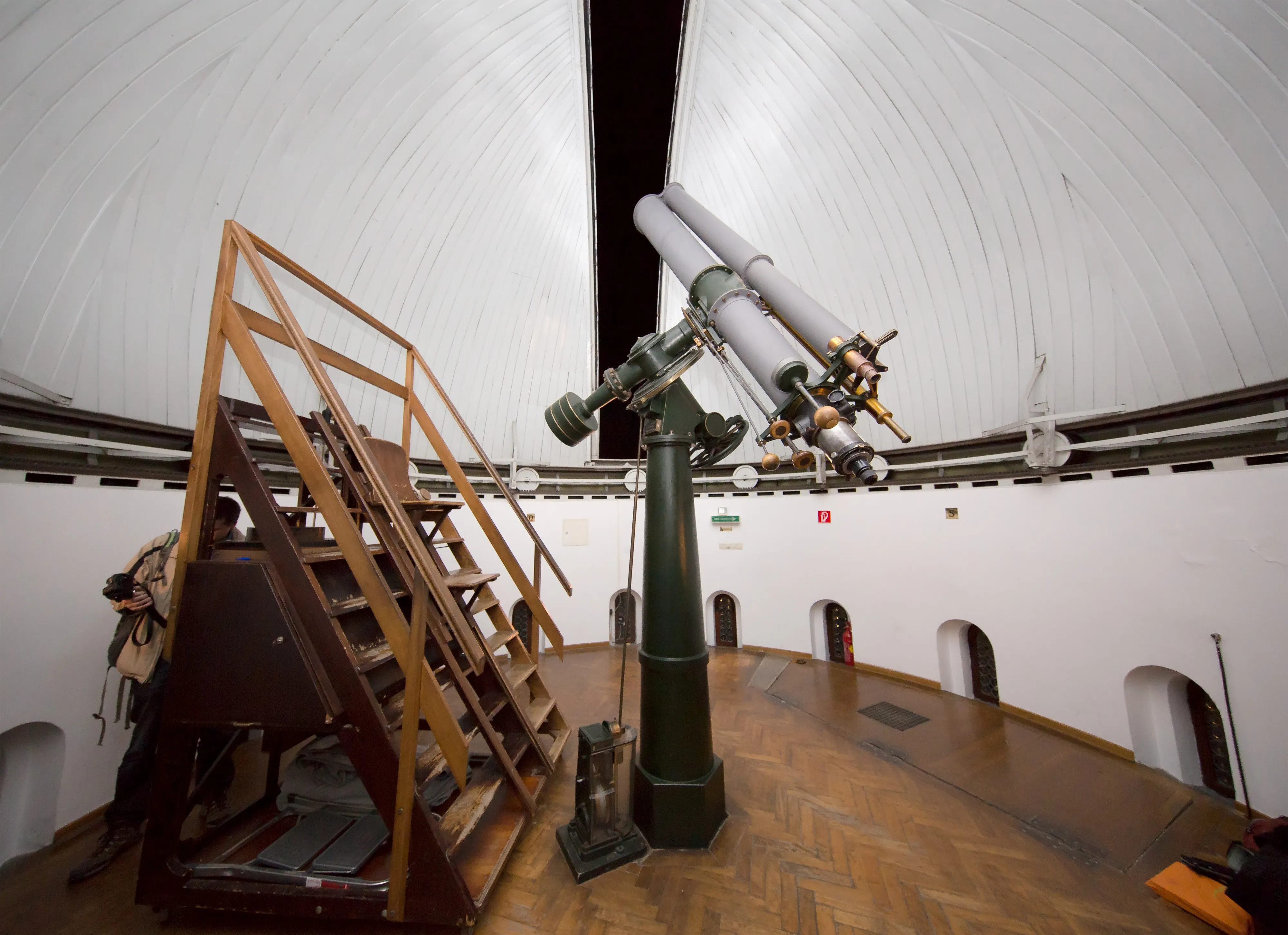 Gran telescopio refractor dentro de una sala de observatorio abovedada con escaleras de madera y un piso pulido