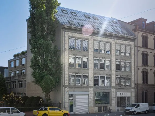 Buitenaanzicht van een gebouw met meerdere verdiepingen en een grote glazen dakconstructie, een hoge boom aan de linkerkant en geparkeerde voertuigen op de straat beneden.