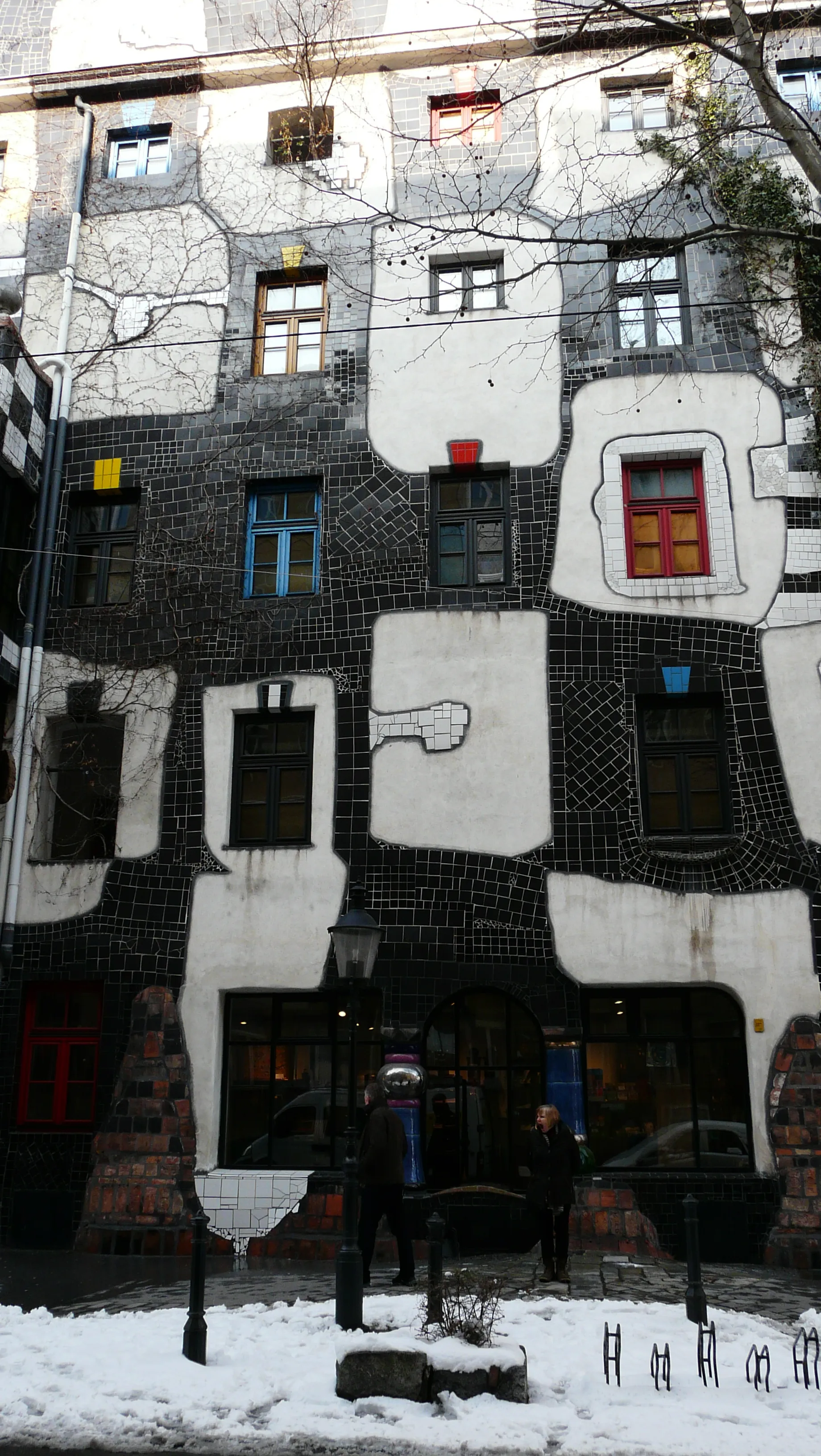 Ośnieżona ulica przed Kunst Haus Wien, budynek o charakterystycznej fasadzie z kolorowymi, nieregularnymi oknami i mozaikowymi wzorami.