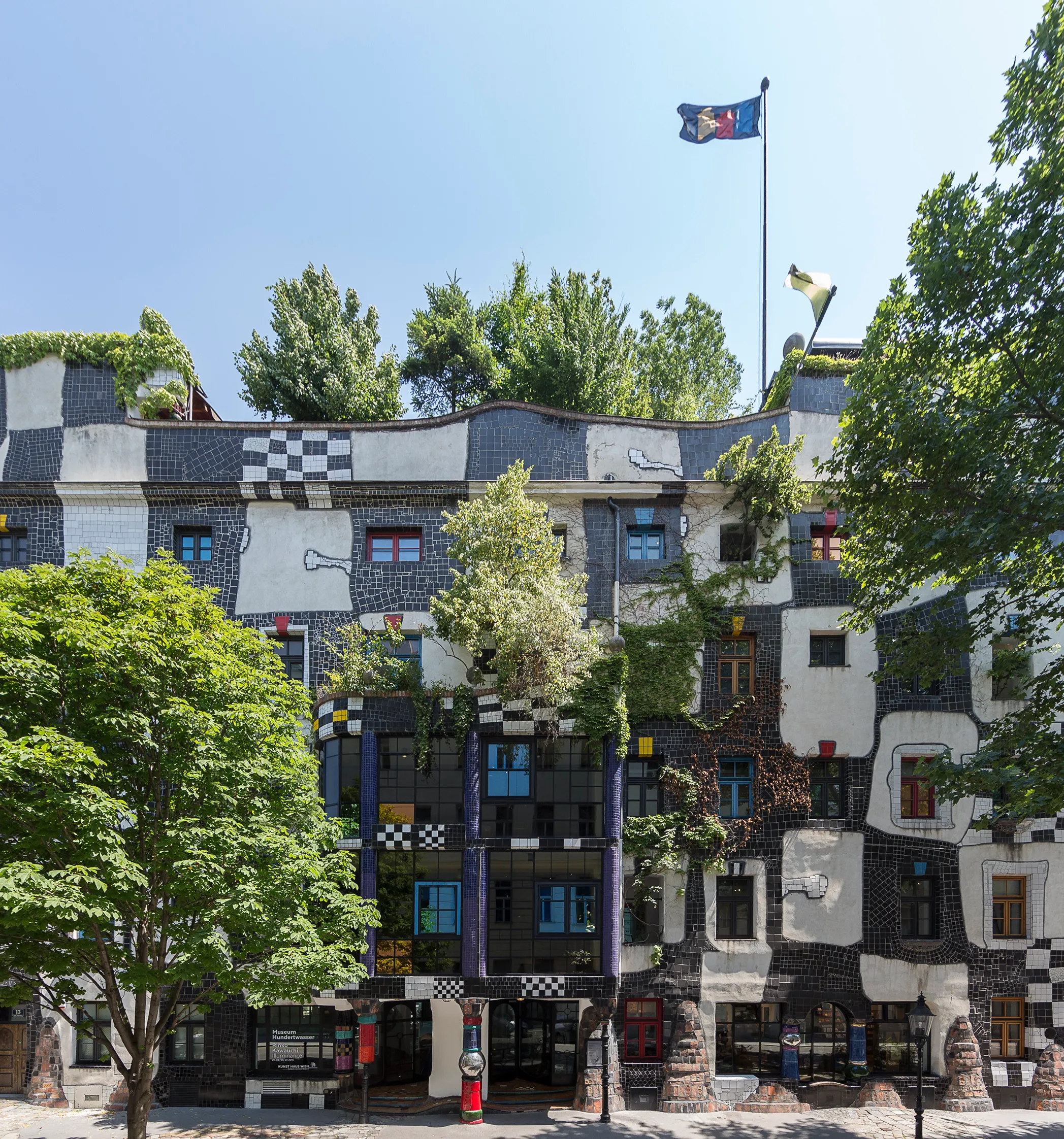 Zewnątrz Kunst Haus Wien z kolorową, nieregularną architekturą, wzorami w kratę i drzewami rosnącymi na fasadzie pod błękitnym niebem