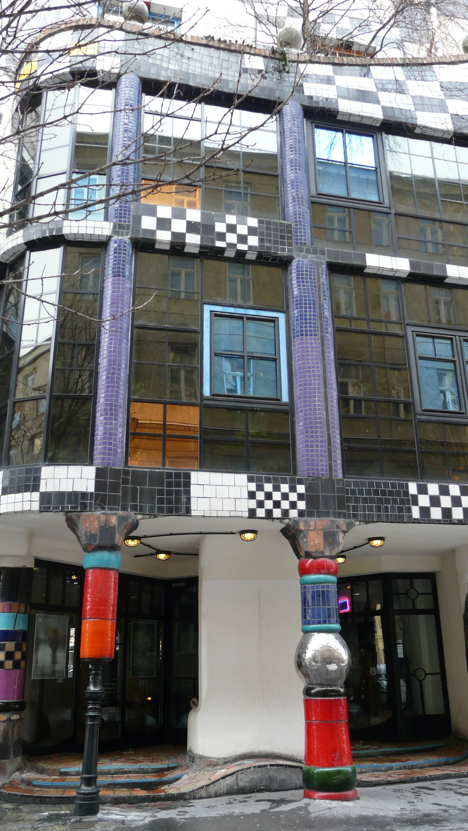 Budynek Kunst Haus Wien z kolorową fasadą pokrytą mozaiką, wzorami w kratę i żywymi, nieregularnymi kolumnami przy wejściu.
