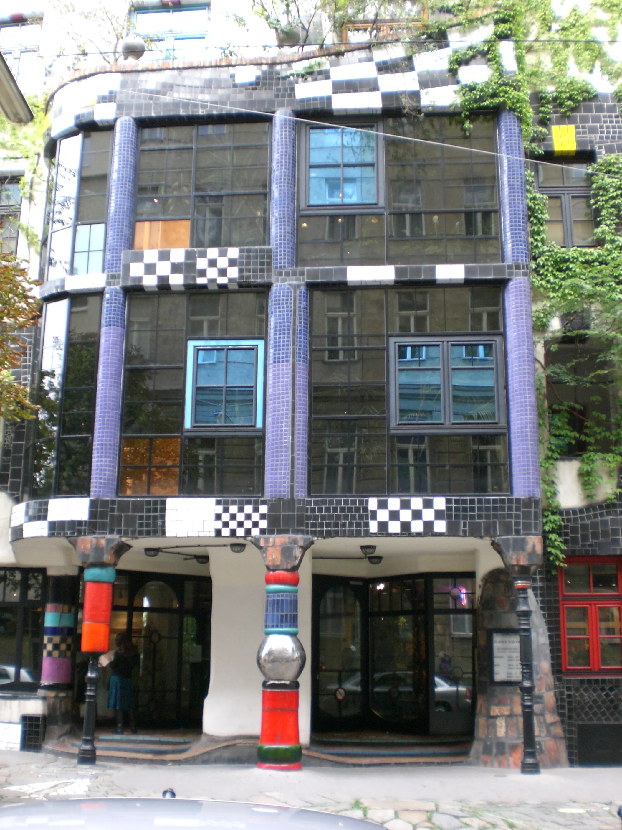 Zewnętrzna część Kunst Haus Wien, budynku z kolorową fasadą pokrytą mozaikowymi płytkami, z wzorami w kratę, fioletowymi kolumnami i nieregularnymi oknami.