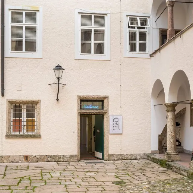 White building facade with arched colonnade, green entrance door, wall-mounted lamp, and cobblestone courtyard at Kunst im Traklhaus