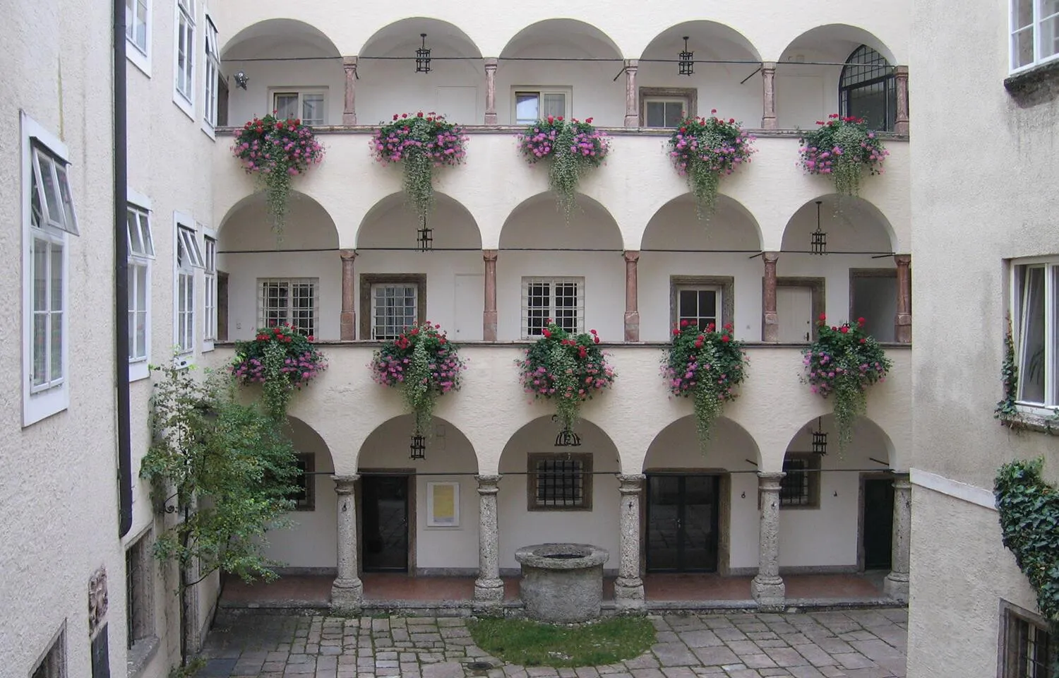 A multi-story courtyard with white arched walkways, hanging flower baskets with purple flowers, and a stone well in the center.