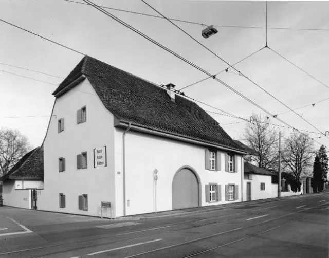 Zwart-wit exterieur van Kunst Raum Riehen, een wit gebouw met een donker schuin dak, een boogvormige deuropening, ramen met luiken en bovenleidingen van de tram.