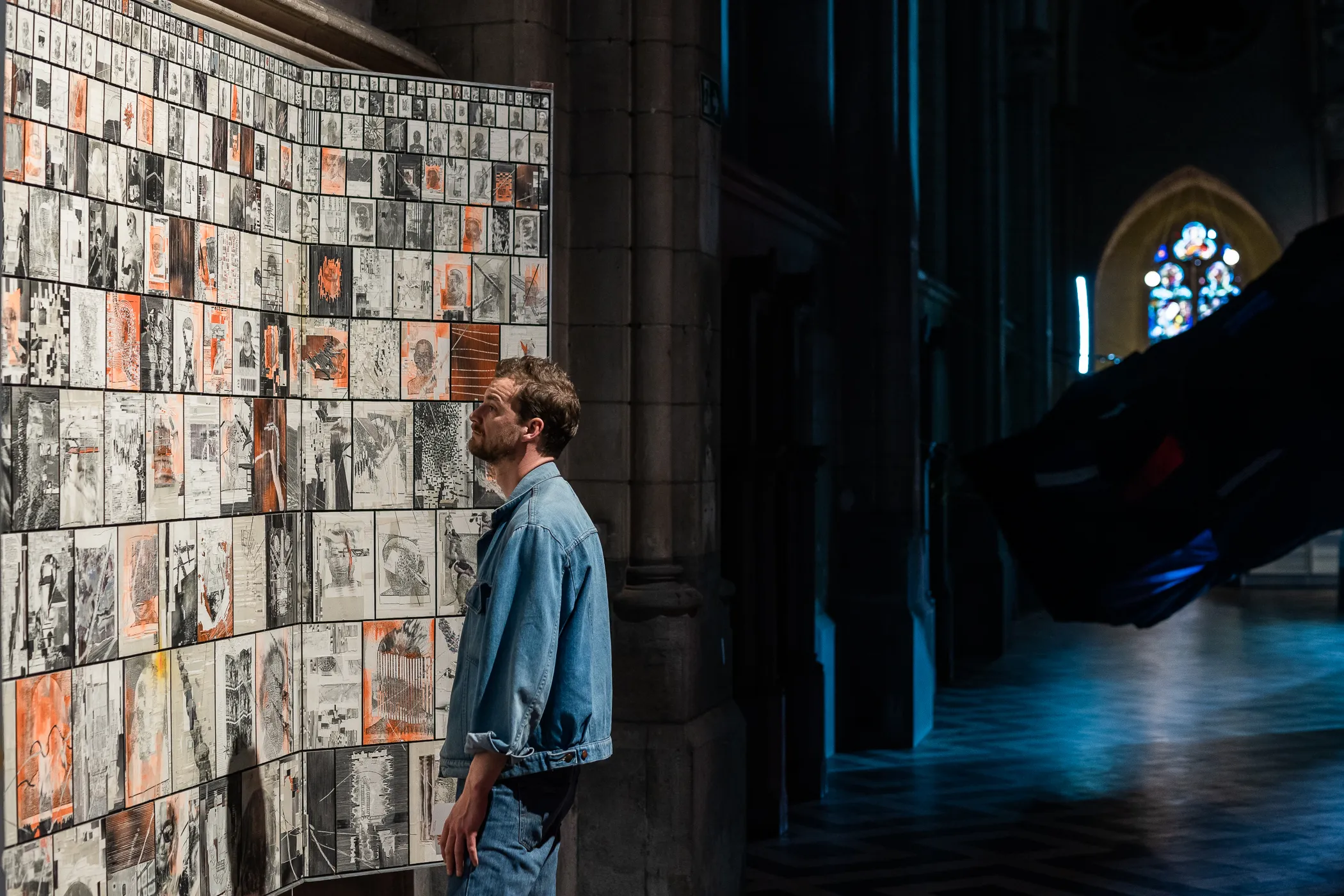 Un hombre con camisa de mezclilla observa una gran instalación mural cubierta de pequeños dibujos dentro de una sala tenuemente iluminada con una vidriera.