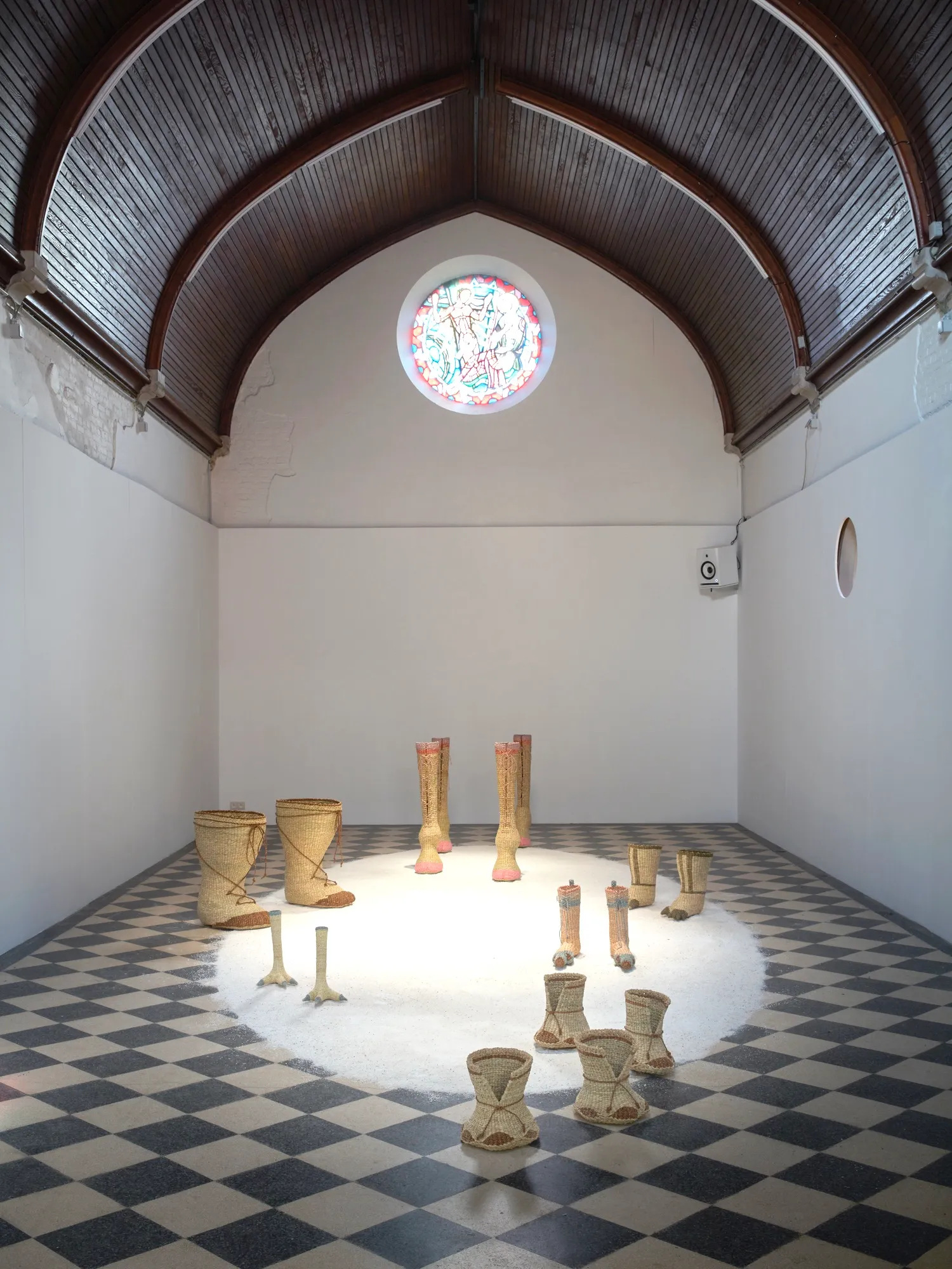 Instalación artística de vasijas de cerámica dispuestas en círculo sobre una alfombra blanca en una sala con suelo de damero y techo abovedado con una vidriera.
