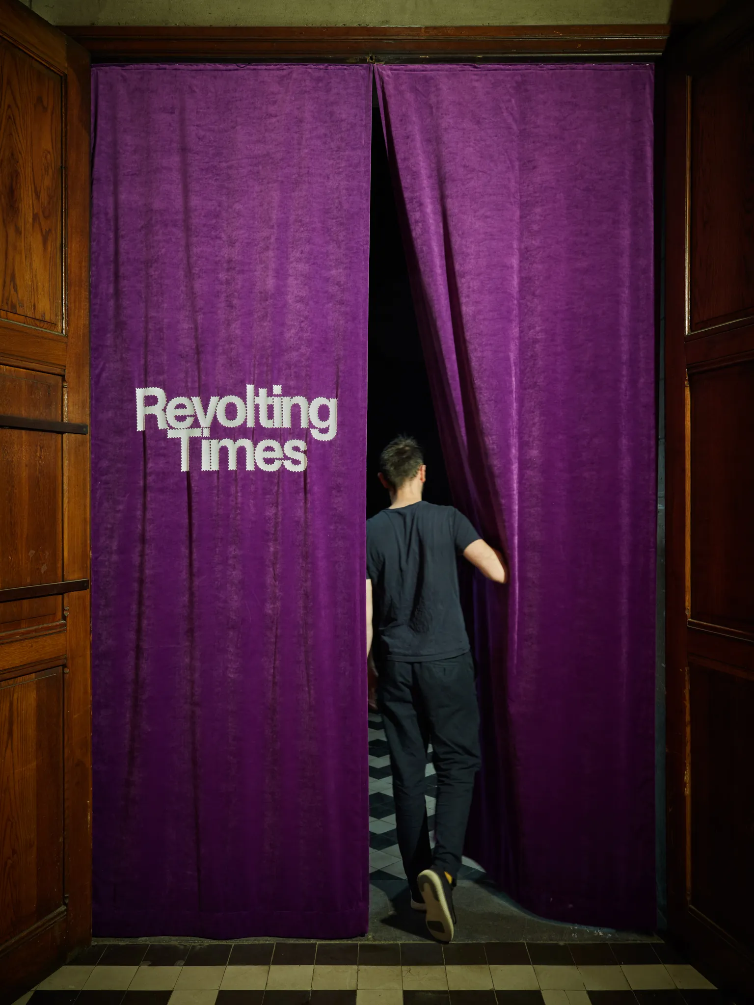Una persona atraviesa unas grandes cortinas de terciopelo morado con el texto en blanco "Revolting Times" en la entrada de una exposición enmarcada por paneles de puertas de madera oscura.
