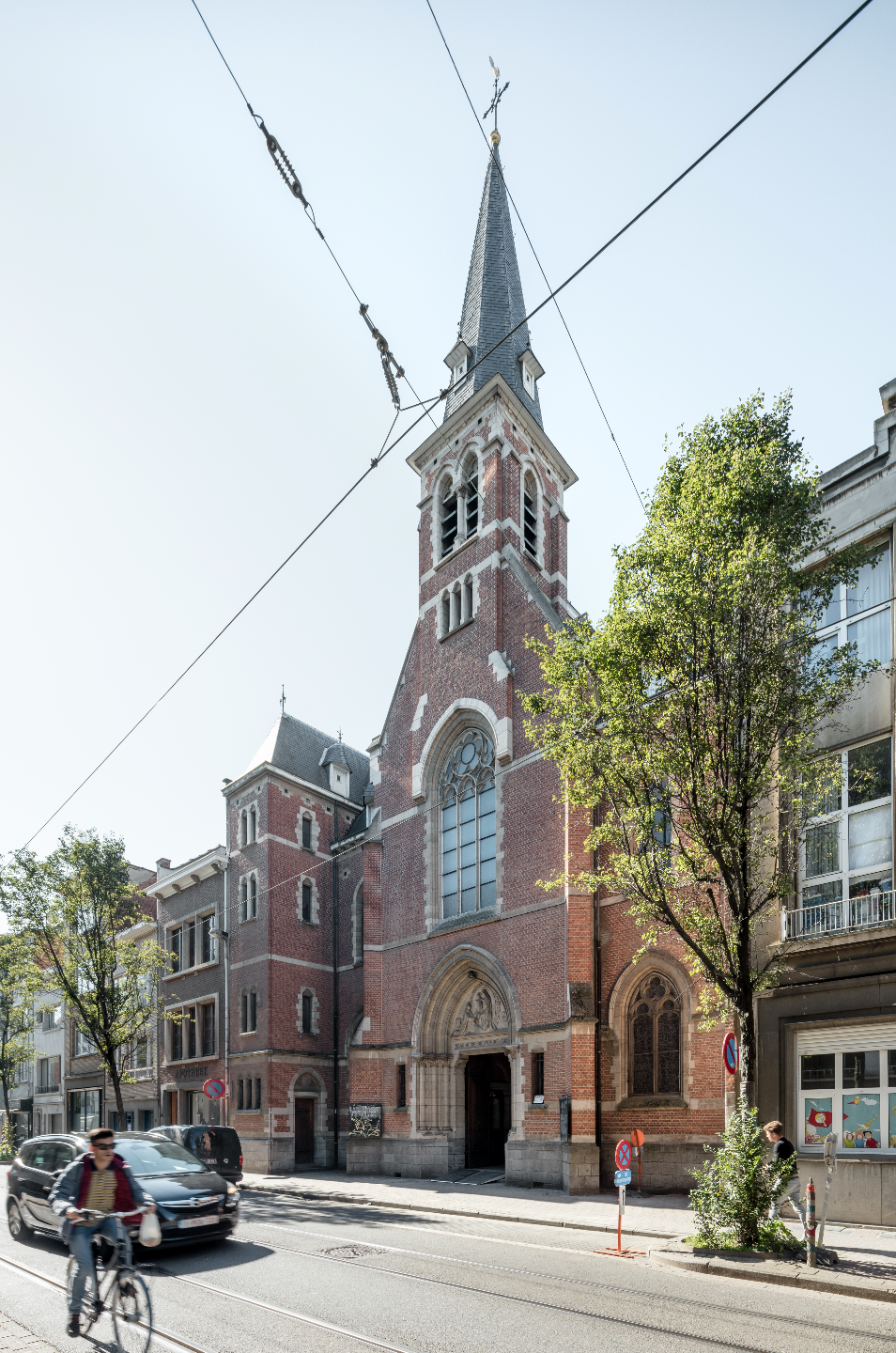 Edificio de iglesia de ladrillo con una torre alta y puntiaguda y una entrada en arco en una calle de la ciudad con líneas de tranvía elevadas.