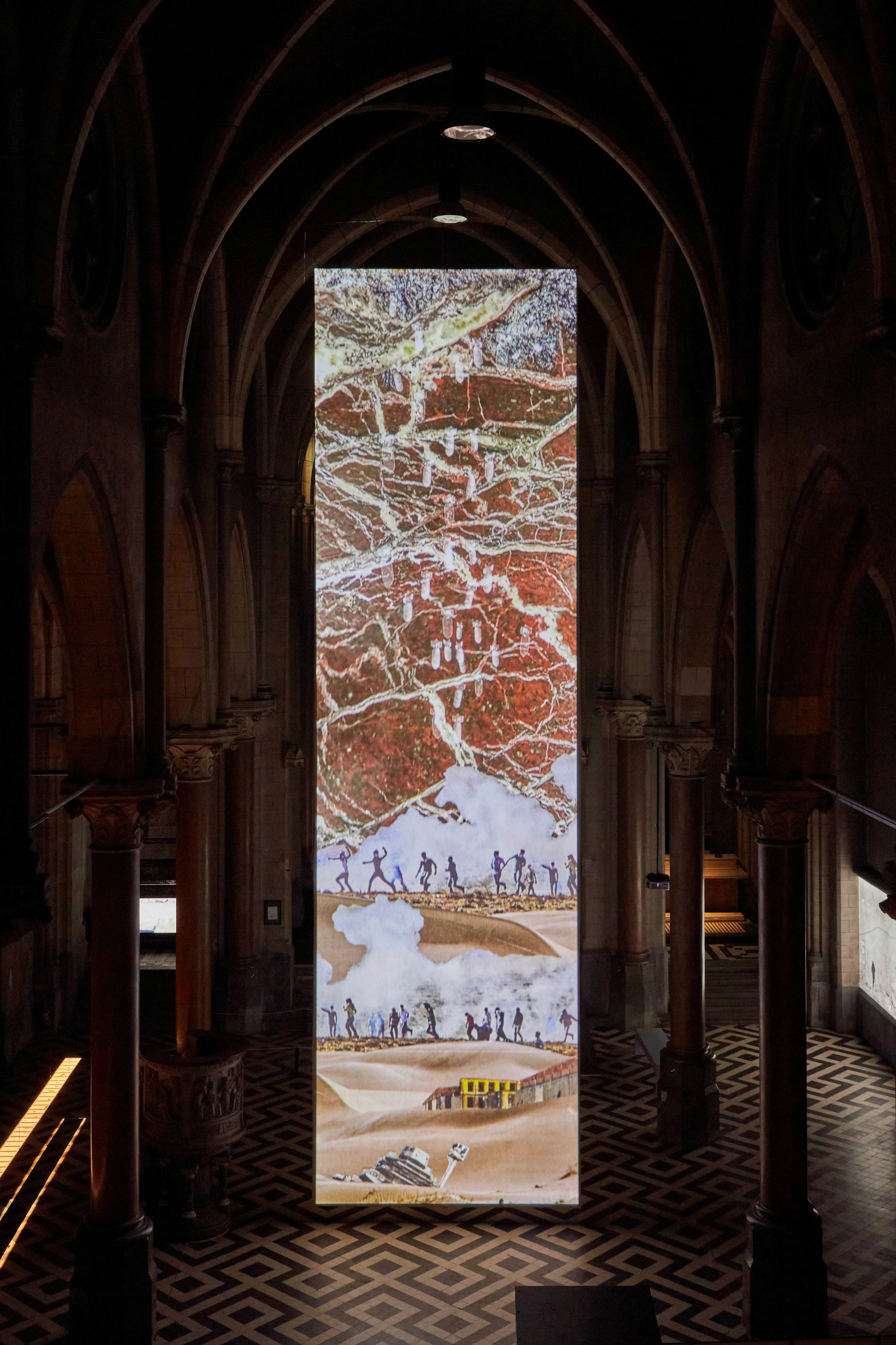 Proyección vertical a gran escala de una obra de arte que representa figuras corriendo a través de un paisaje, expuesta en la nave de un antiguo monasterio con arcos góticos